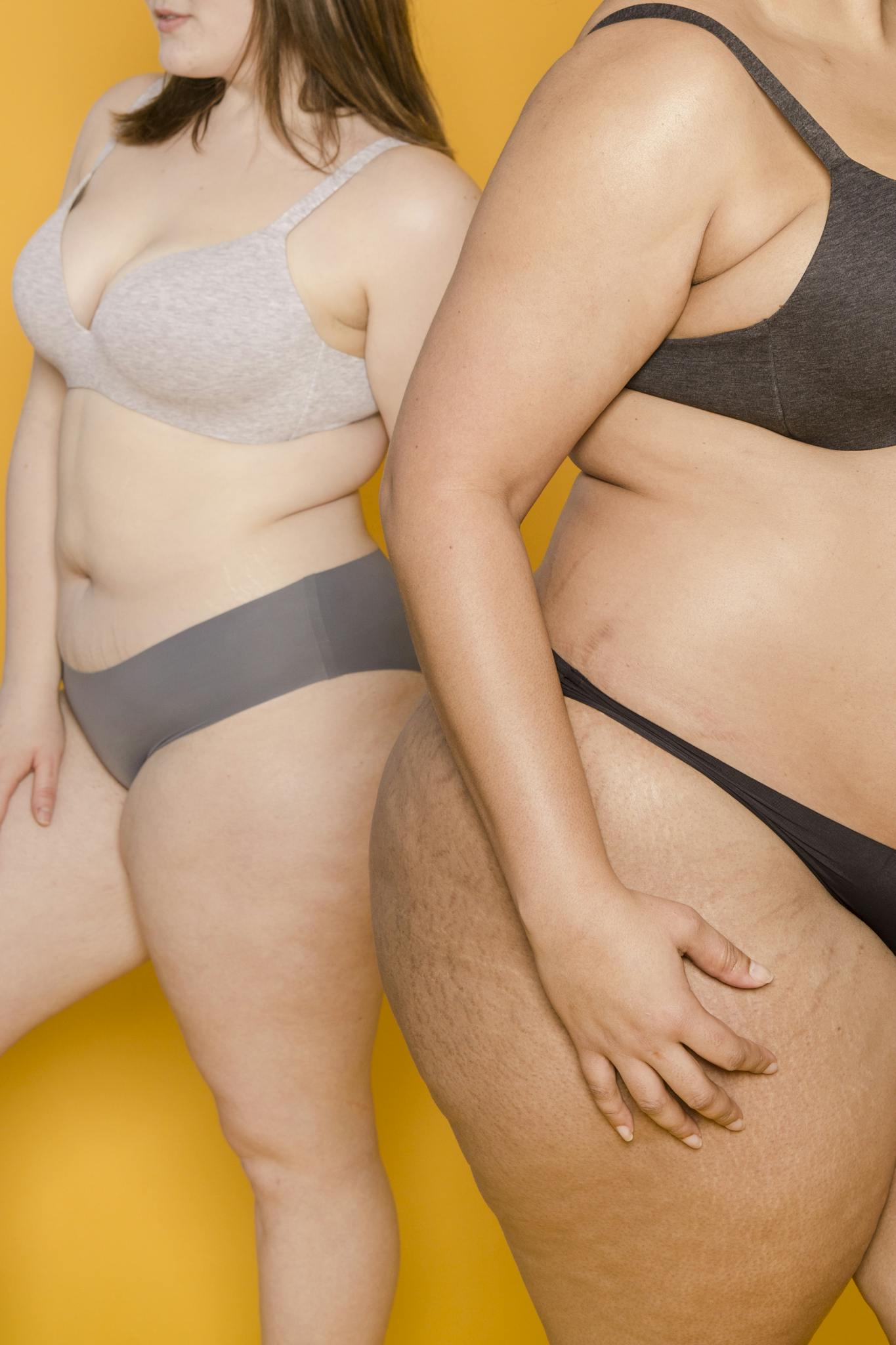 Two women confidently pose in lingerie against a yellow background, promoting body positivity.
